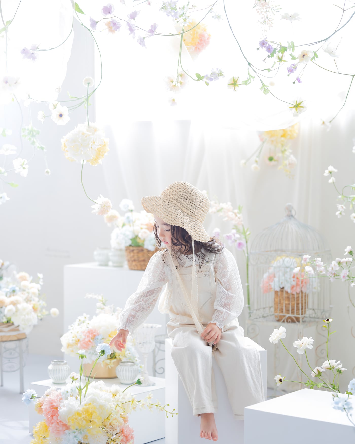 淡い色の花に囲まれた白い空間で、麦わら帽子をかぶって座る女の子(岡崎・岡崎スタジオ)