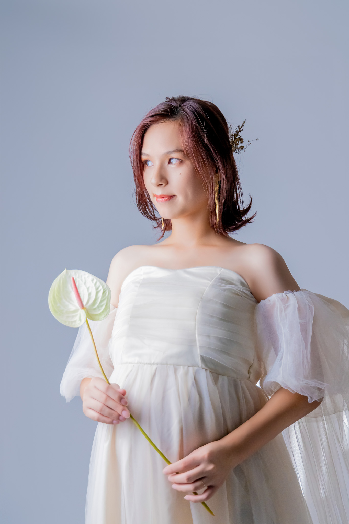 オフショルダーの白いチュールドレスをまとい、白いアンスリウムを手に横を向く妊婦