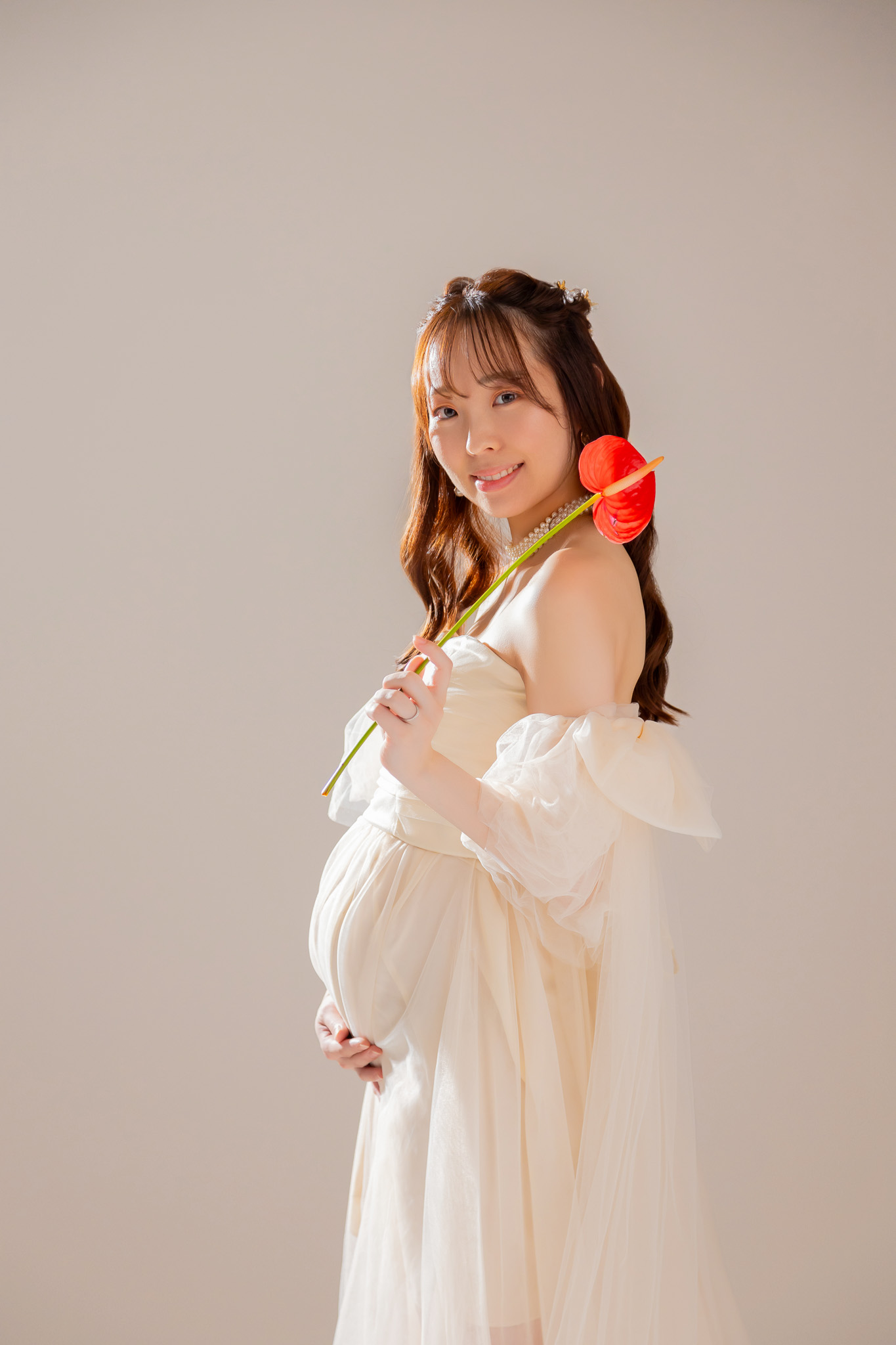 窓からの光を生かしたスタジオで、赤い花を手にして微笑む妊婦