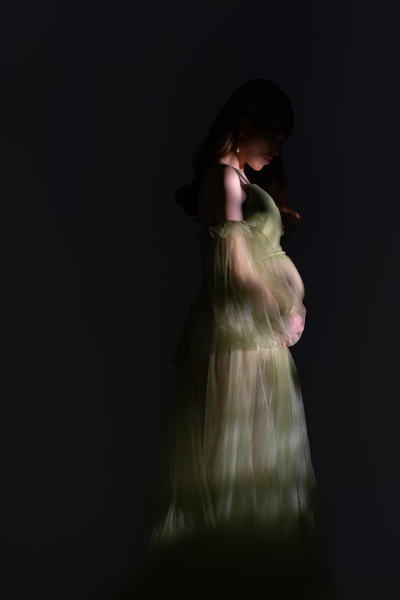 暗い背景の中、淡いグリーンのチュールドレスでお腹に手を添え、うつむく妊婦
