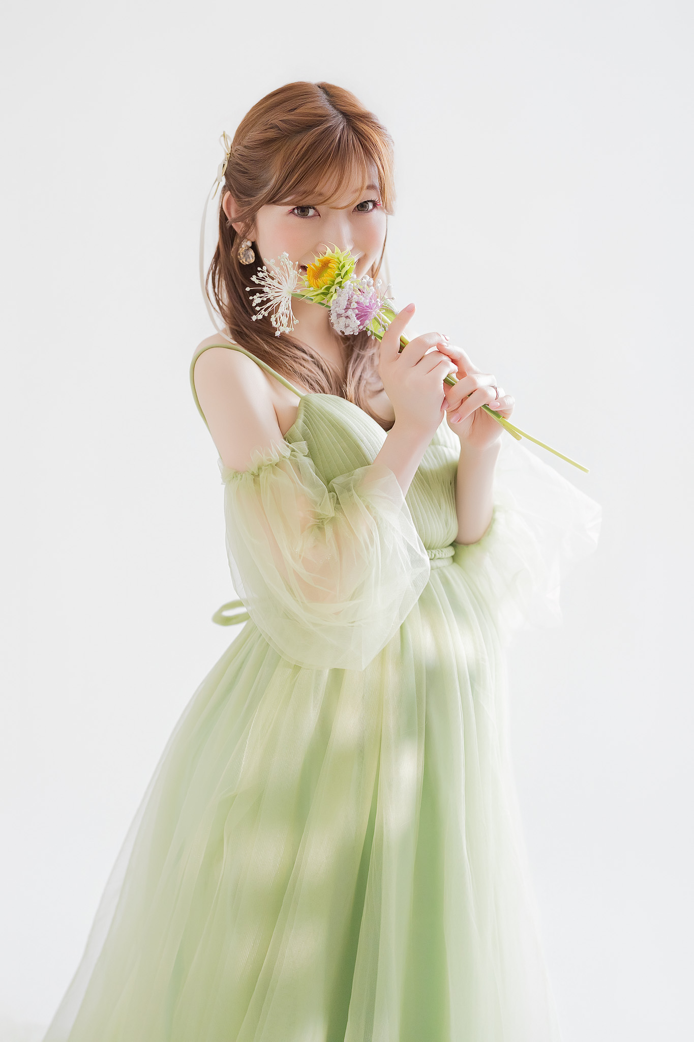 胸の下で切り替わったグリーンのチュールドレスをまとい、小花を手にカメラを見る妊婦