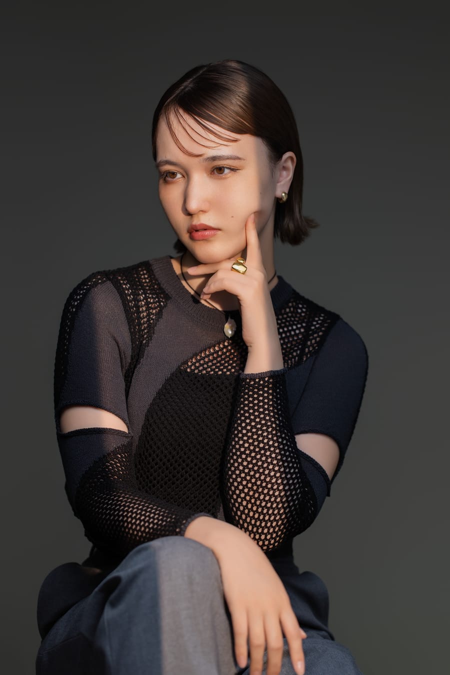 濃いグレーの背景で椅子に腰かけ、頬に手を添えた黒い装いのプロフィール写真